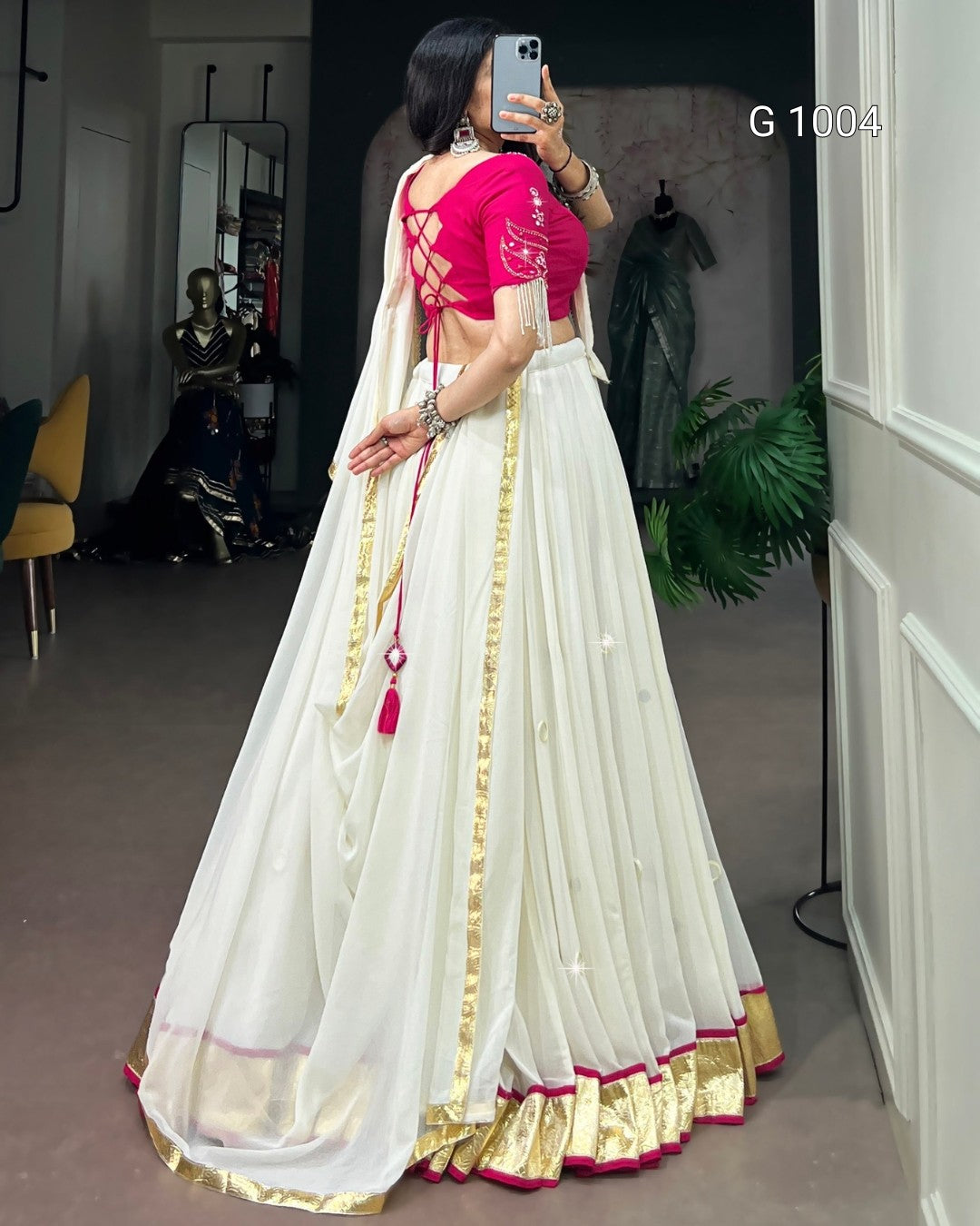 A woman takes a mirror selfie in a room wearing a traditional Indian outfit consisting of a white flared skirt (lehenga) with gold zari borders and a bright pink embroidered blouse. The text 'G 1004' is visible in the top right corner.