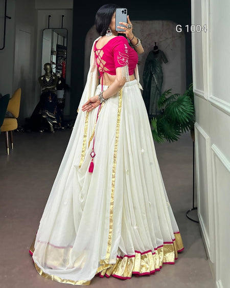 A woman takes a mirror selfie in a room wearing a traditional Indian outfit consisting of a white flared skirt (lehenga) with gold zari borders and a bright pink embroidered blouse. The text 'G 1004' is visible in the top right corner.