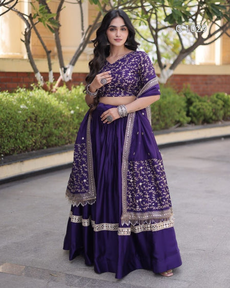 A woman modeling a traditional Indian outfit consisting of a deep purple lehenga skirt with intricate silver embroidery, a matching embroidered purple blouse, and a coordinating dupatta, set against an outdoor background of greenery and a building.