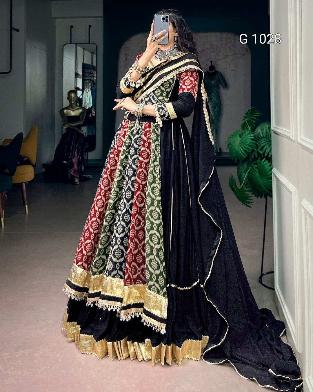 A woman takes a mirror selfie wearing a traditional Indian outfit consisting of a black dupatta, a heavily embroidered black lehenga, and a red and green patterned blouse. The outfit features intricate gold and silver embroidery and borders.