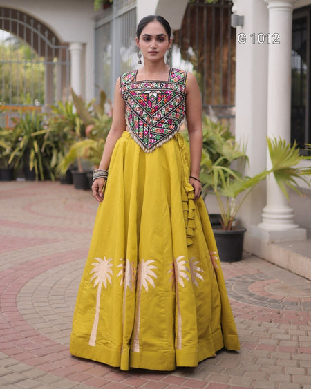 A woman modeling a traditional Indian anarkali gown in mustard yellow, featuring a heavily embroidered bodice with pink, green, and silver details, and cream-colored palm tree motifs near the hem, set against an outdoor courtyard background.