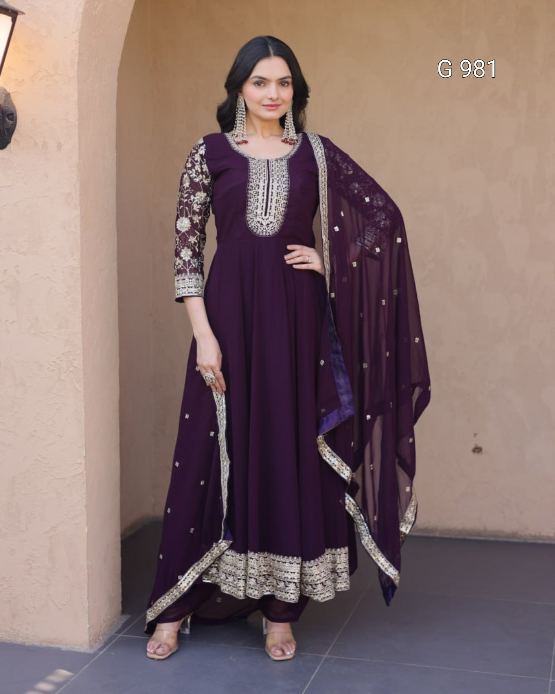 A woman models a deep purple traditional Indian salwar kameez suit with intricate silver embroidery on the sleeves, neckline, and hem, paired with a matching sheer dupatta. She is standing in front of a beige stucco wall.