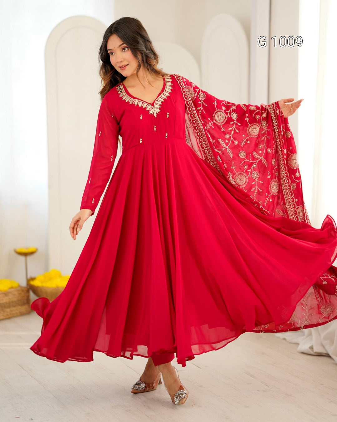 A woman modeling a bright red long-sleeve Anarkali gown with gold embroidery and a matching sheer red dupatta, posed in a light-colored indoor setting.