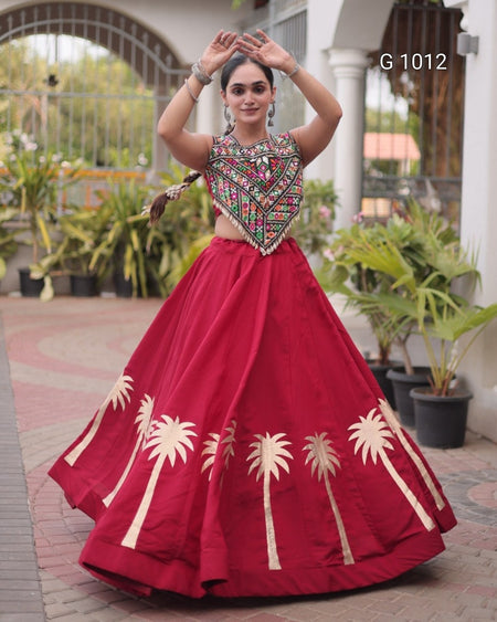 A woman modeling a traditional Indian outfit featuring a deep red flared skirt with gold palm tree embroidery and a heavily embellished embroidered crop top, set against an outdoor patio background with potted plants.