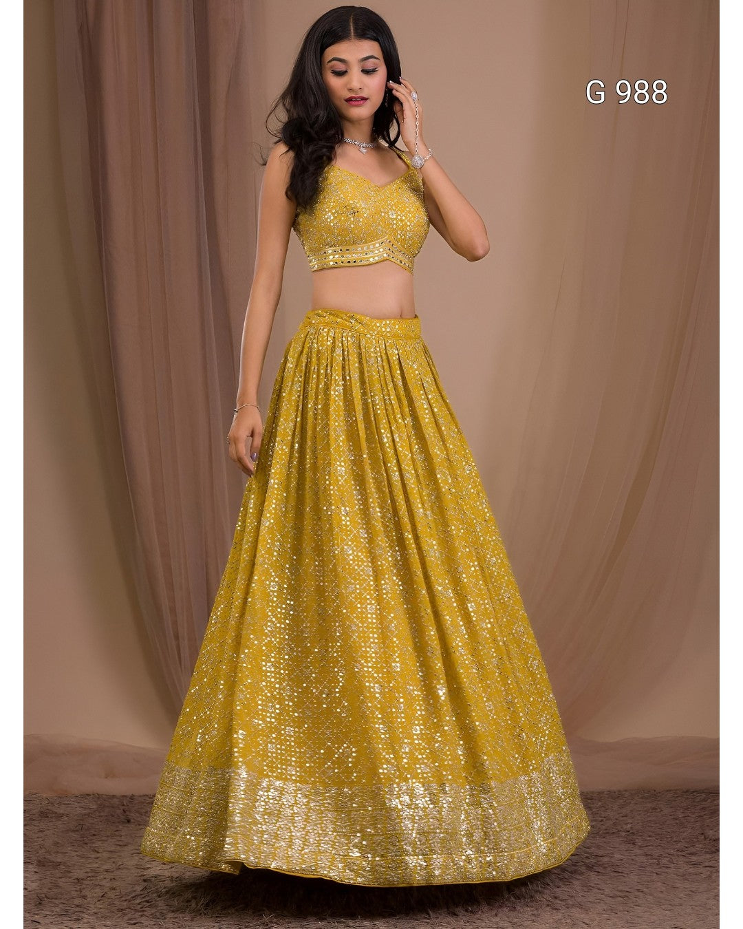 A woman modeling a bright yellow embellished lehenga choli set against a draped beige studio backdrop. The set includes a sleeveless crop top and a full flared skirt with sequined detailing and a wide border hem.