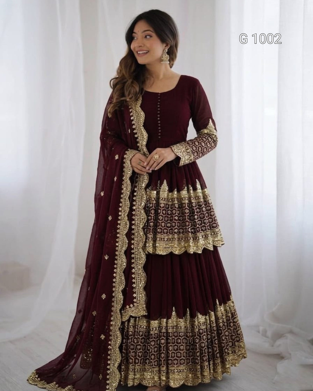 A woman modeling a traditional maroon Anarkali gown with intricate gold embroidery and a matching dupatta, set against a soft white curtain backdrop.