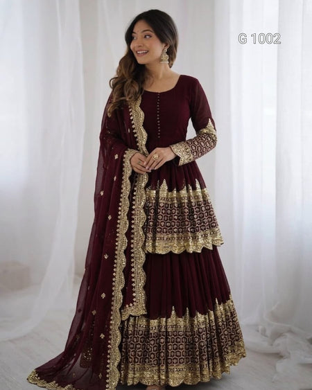 A woman modeling a traditional maroon Anarkali gown with intricate gold embroidery and a matching dupatta, set against a soft white curtain backdrop.