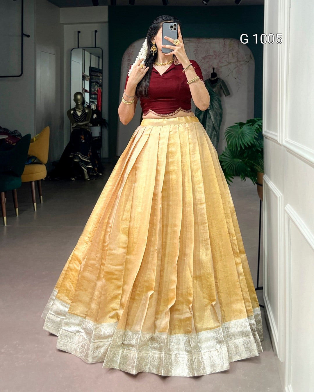 A woman takes a mirror selfie in a boutique wearing a traditional Indian outfit consisting of a deep maroon velvet blouse and a voluminous golden-yellow silk skirt with a silver border. The boutique interior is visible in the background.