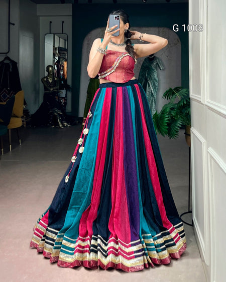 A woman takes a mirror selfie wearing a traditional Indian lehenga choli outfit. The choli is a dusty rose pink velvet with a silver beaded border, and the flared lehenga features vertical stripes of teal blue, magenta pink, and navy blue, finished with a striped border at the hem.