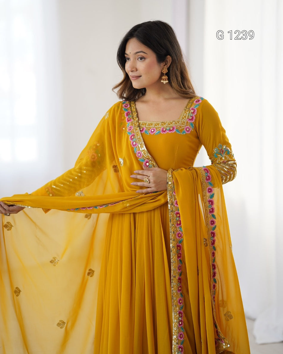 A woman modeling a mustard yellow traditional outfit with colorful embroidered borders, holding out a matching sheer dupatta in a bright indoor setting.