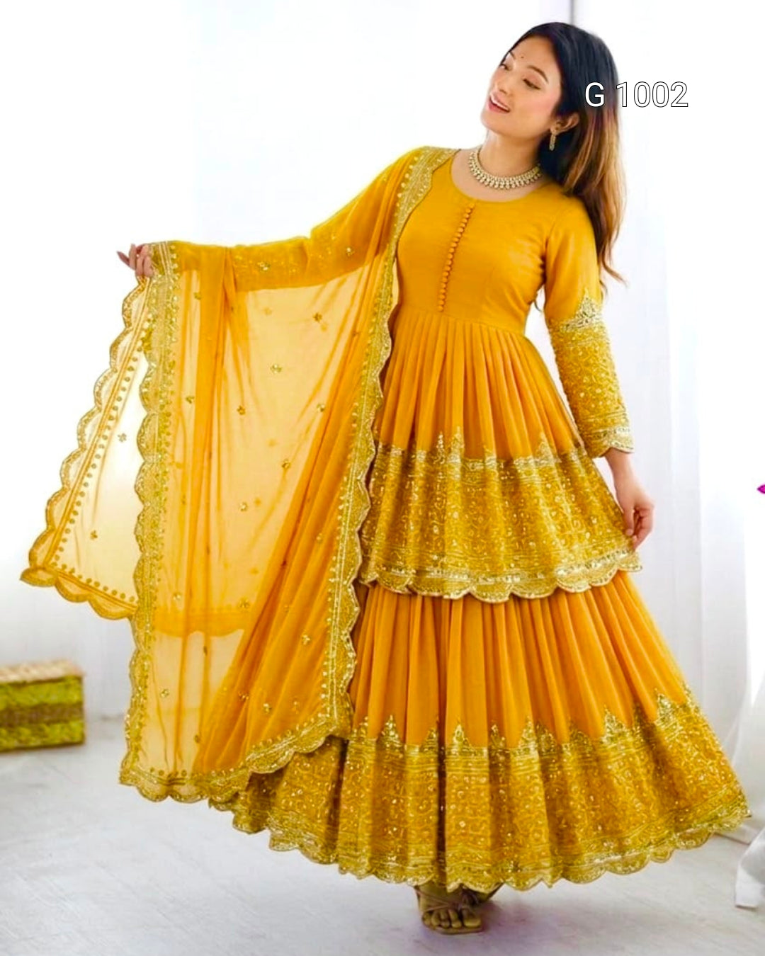 A woman modeling a bright yellow traditional Anarkali gown with a matching sheer dupatta, featuring intricate gold embroidery and scalloped borders. The image has a white background with a small text overlay 'G 1002' in the top right.