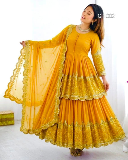 A woman modeling a bright yellow traditional Anarkali gown with a matching sheer dupatta, featuring intricate gold embroidery and scalloped borders. The image has a white background with a small text overlay 'G 1002' in the top right.