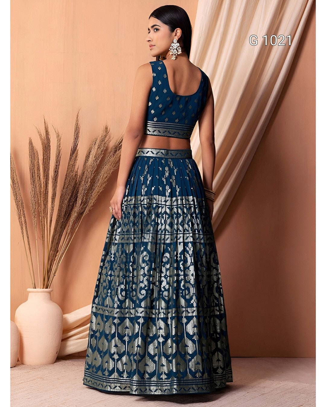 A woman modeling a dark teal blue and silver lehenga choli set, shown from the back. The set features intricate silver mirror-work patterns on a dark teal fabric, with a sleeveless crop top and a full flared skirt.