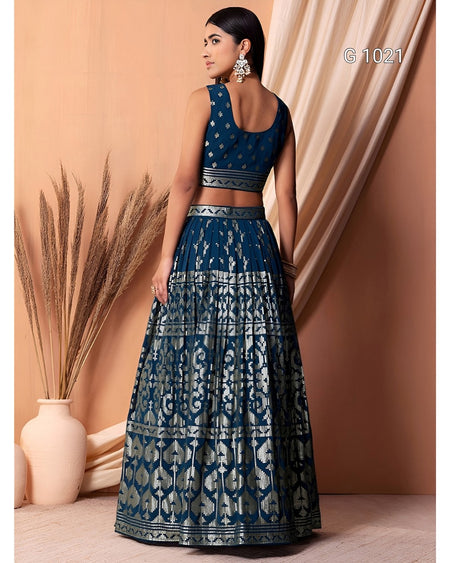 A woman modeling a dark teal blue and silver lehenga choli set, shown from the back. The set features intricate silver mirror-work patterns on a dark teal fabric, with a sleeveless crop top and a full flared skirt.