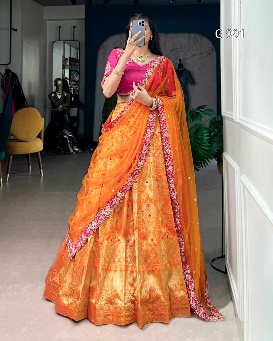 A woman takes a mirror selfie wearing a traditional Indian saree. The saree features a bright orange body with intricate gold zari embroidery, a pink blouse, and a matching orange dupatta with a pink floral border.