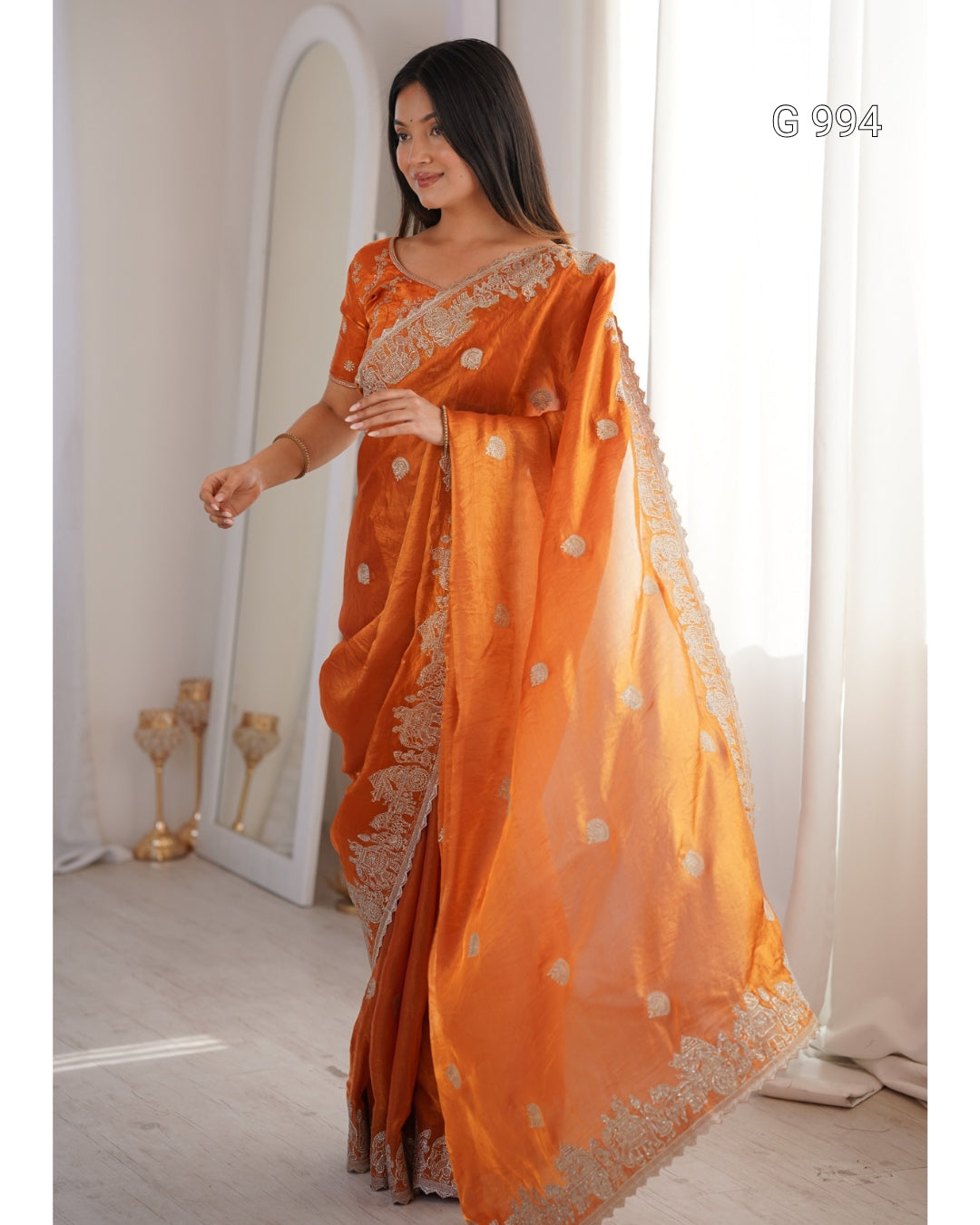 A woman modeling an orange saree with gold floral motifs and a gold embroidered border, standing in a bright indoor setting with a mirror and white curtains.