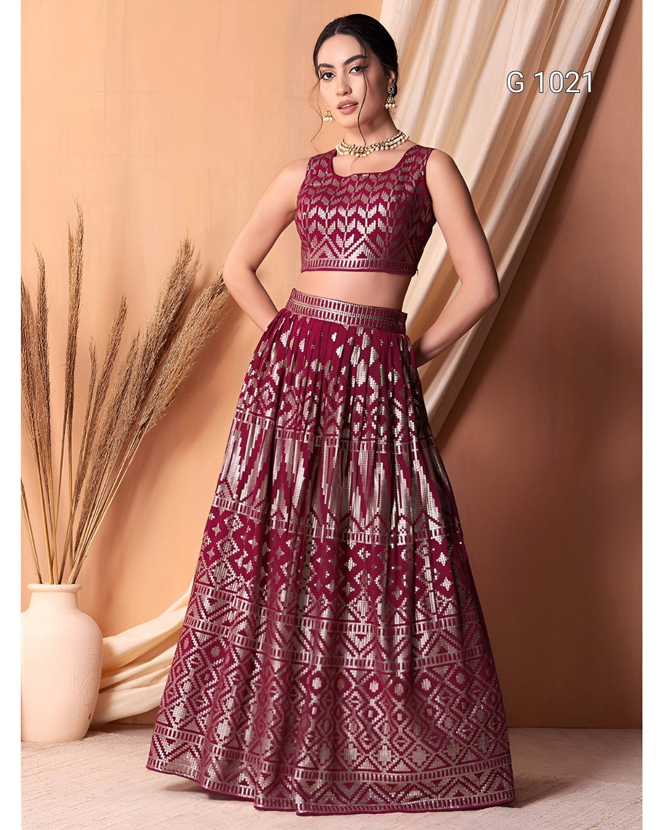 A woman models a deep magenta lehenga choli set with intricate silver geometric embroidery, standing in front of a warm beige draped backdrop.