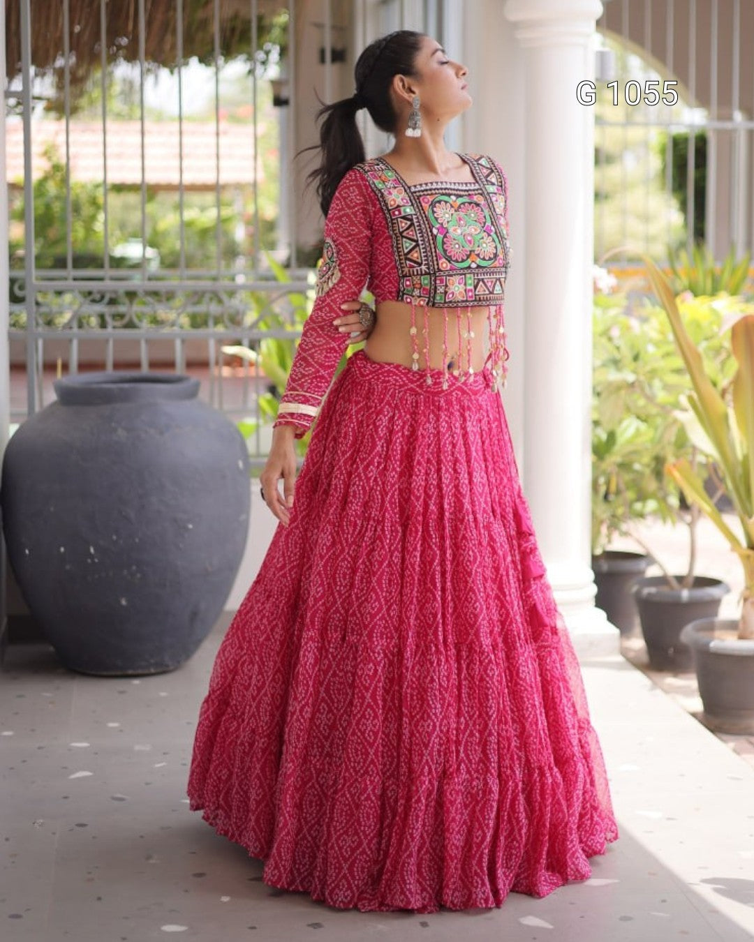 A woman wearing a bright pink embroidered traditional outfit with a flared skirt and patterned blouse, standing on a porch with potted plants and white columns in the background.