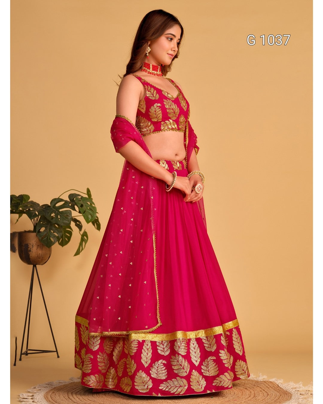 A woman models a bright pink lehenga choli set with gold leaf embroidery and gold dot patterns, standing against a beige background with a small potted plant on the left.