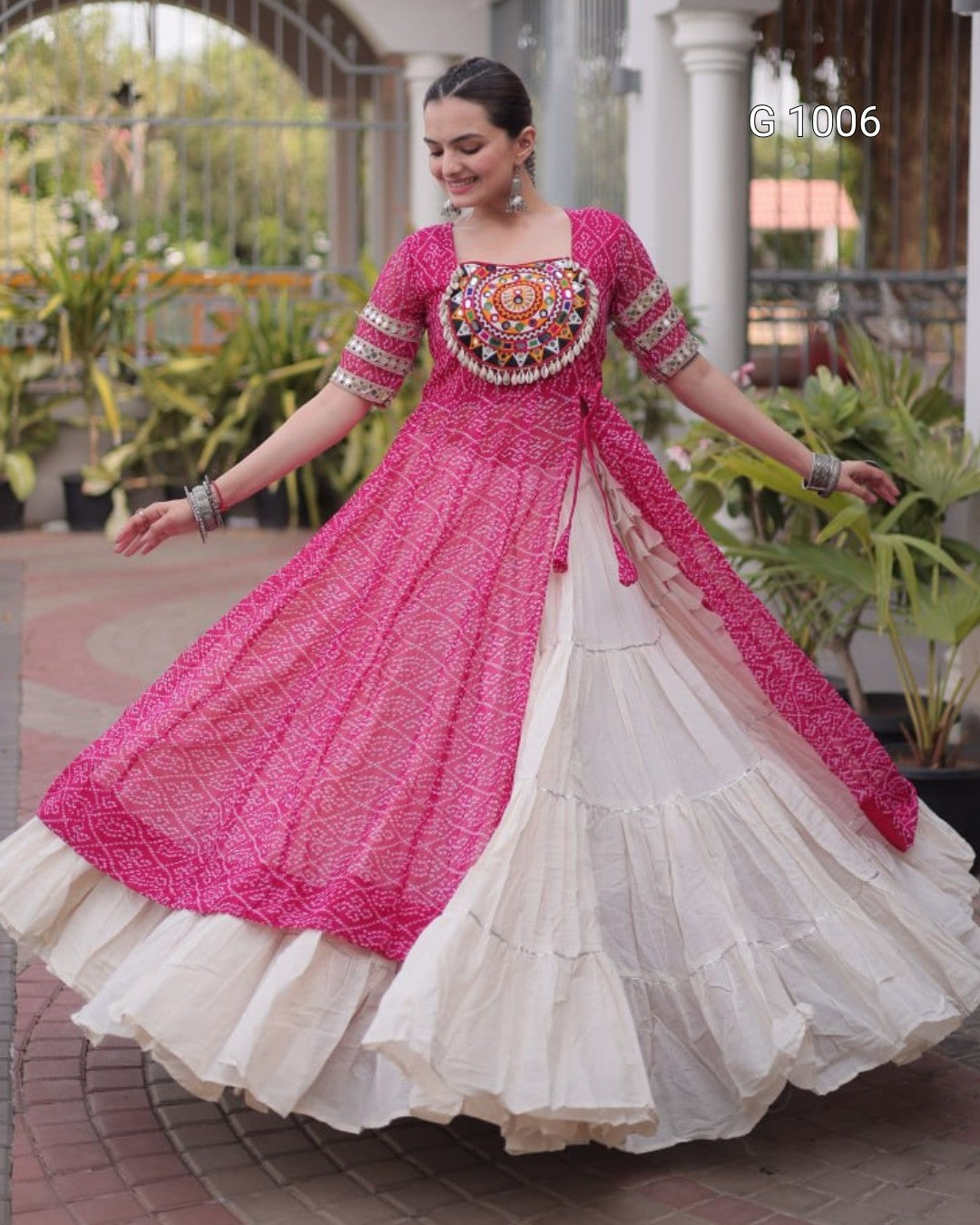 A woman wearing a traditional Indian salwar kameez outfit with a bright pink embroidered kurta and a tiered off-white skirt, posing outdoors in front of a building with columns and plants.