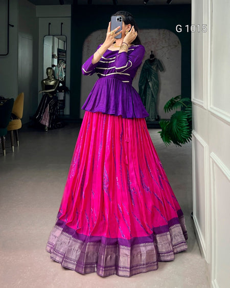 A woman takes a mirror selfie wearing a traditional Indian outfit consisting of a purple peplum top and a bright pink flared skirt with purple borders. The setting appears to be a boutique or dressing room with clothing displays in the background.