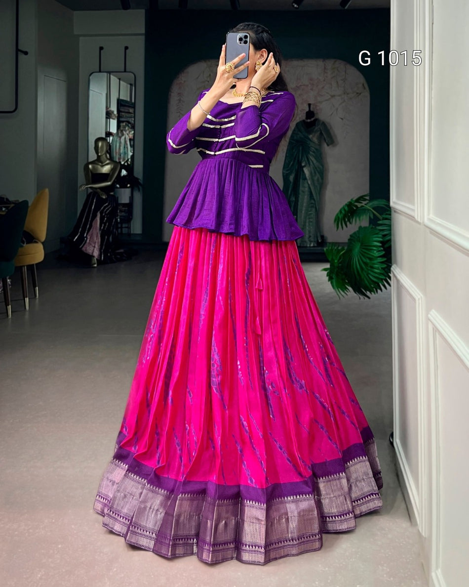 A woman takes a mirror selfie wearing a traditional Indian outfit consisting of a purple peplum top and a bright pink flared skirt with purple borders. The setting appears to be a boutique or dressing room with clothing displays in the background.