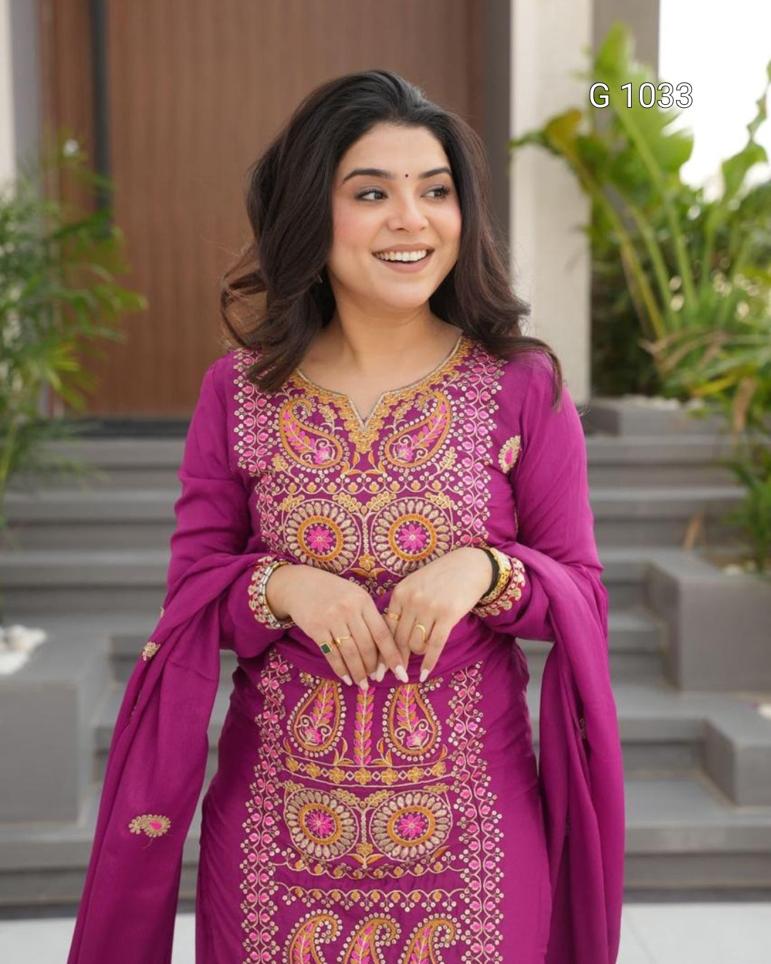 A woman wearing a magenta pink ethnic salwar kameez suit with intricate gold and orange embroidery, standing in front of a modern building with steps and green plants.