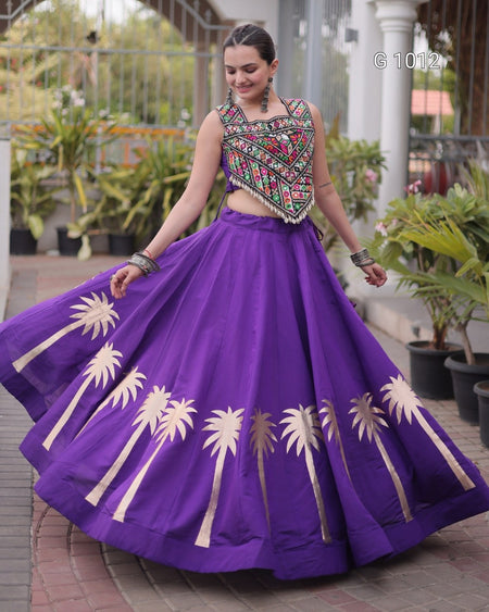 A woman modeling a purple traditional Indian lehenga choli set with embroidered details on the bodice and gold palm tree prints on the skirt, set against an outdoor garden background.