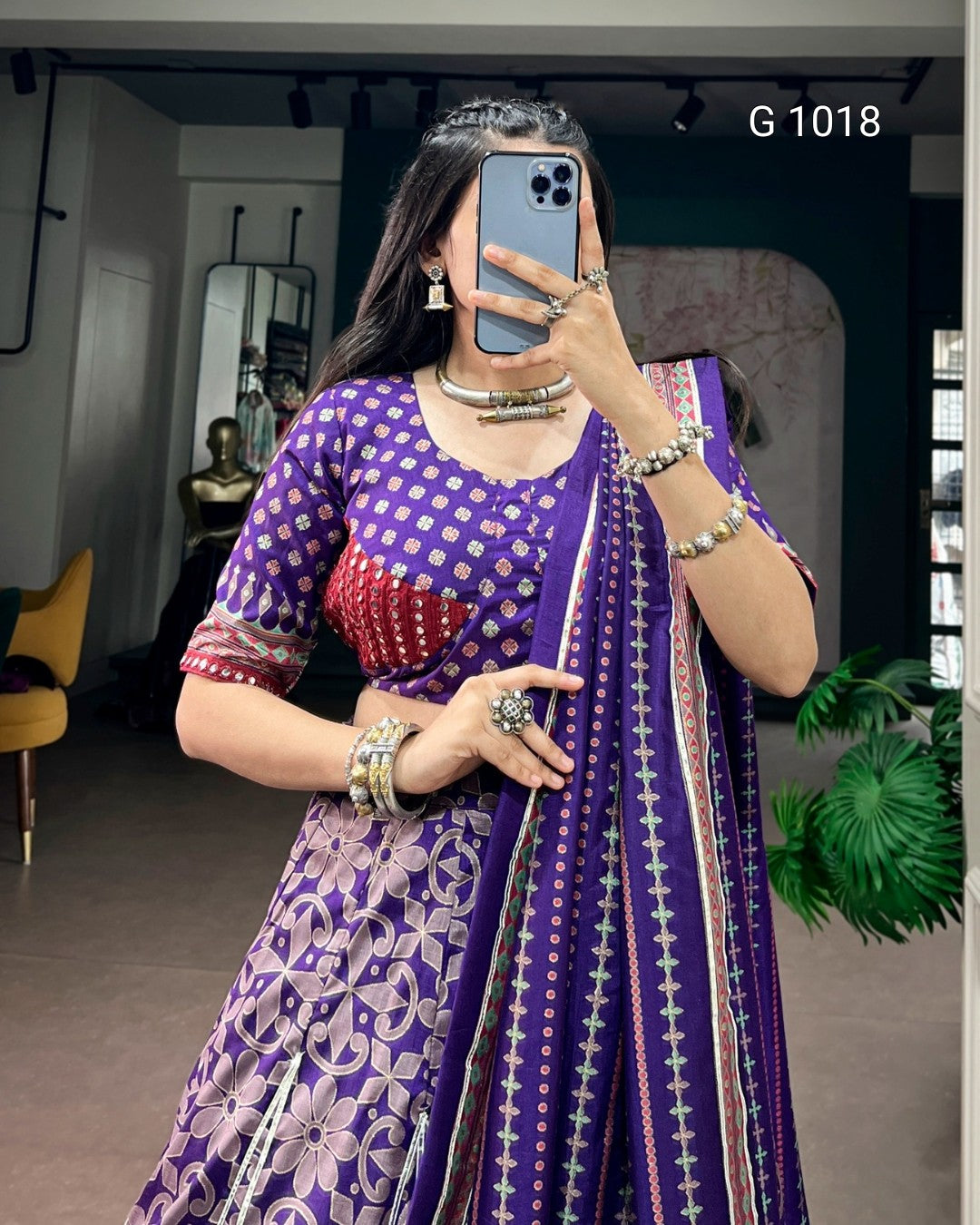 A woman takes a mirror selfie wearing a purple traditional outfit with white floral and geometric patterns, a red embroidered bodice detail, and a matching patterned dupatta. She is wearing multiple silver-toned bangles and rings, and the background shows a modern indoor space with a yellow chair and green plant.