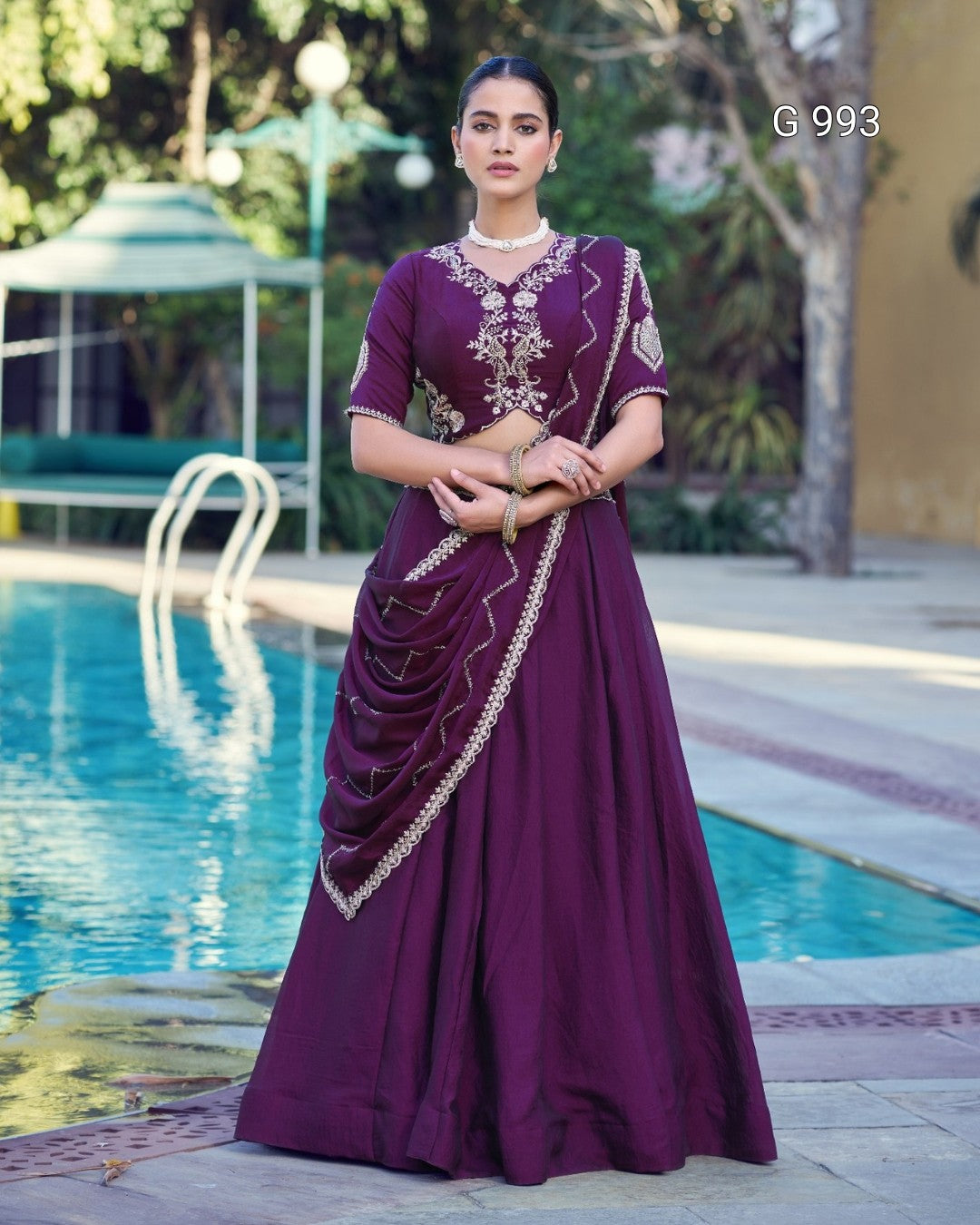 A woman stands by a swimming pool wearing a deep purple traditional dress with gold embroidery and a draped sari. The background shows a pool, trees, and patio seating.