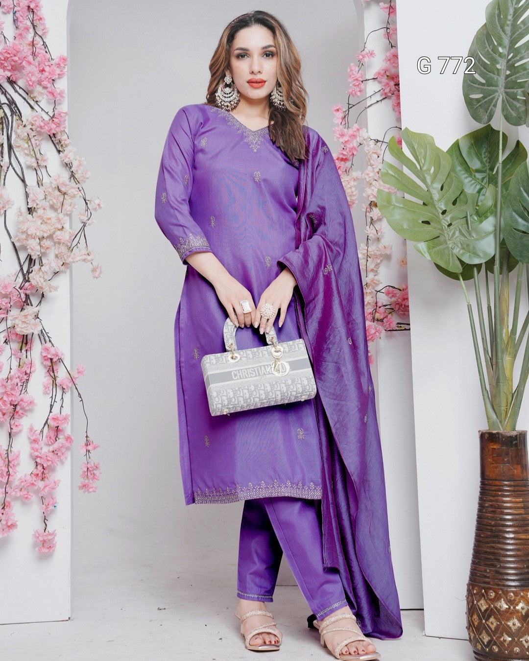 Woman in a purple outfit holding a silver handbag against a white wall with decorative elements.
