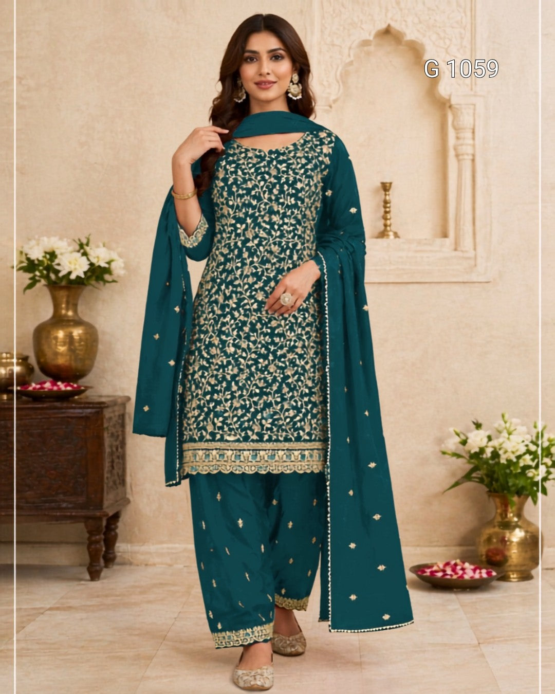 A woman models a teal green ethnic salwar kameez suit with intricate gold floral embroidery, matching pants, and a dupatta, set against a warm beige architectural background with decorative pillars and flower vases.