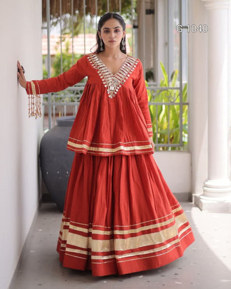 A woman models a traditional Indian red anarkali-style dress with beige and gold striped borders and silver embellishments on the V-neckline, set against a modern outdoor corridor background.