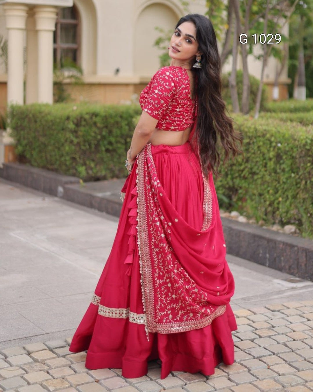 A woman wearing a bright pink traditional Indian lehenga choli set with gold embroidery, standing outdoors on a paved walkway in front of a building with green hedges.