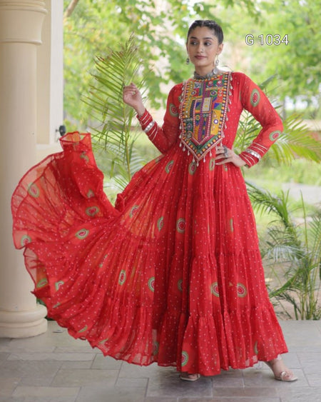 A woman models a vibrant red long-sleeve Anarkali gown with intricate multicolor embroidery on the chest and scattered gold motifs on the flowing skirt, set against a garden background with a pillar.