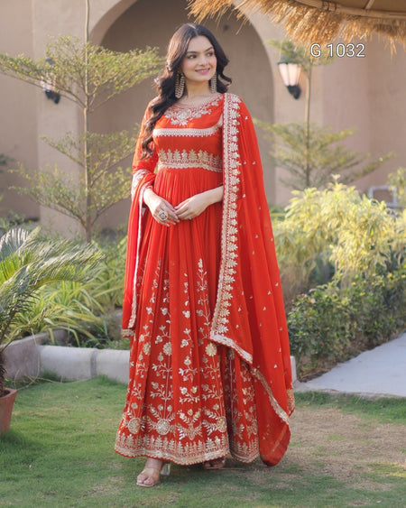 A woman stands outdoors in a garden wearing a bright red traditional outfit with extensive gold embroidery and a matching embroidered dupatta. The background shows a beige building with arches, plants, and a pathway.