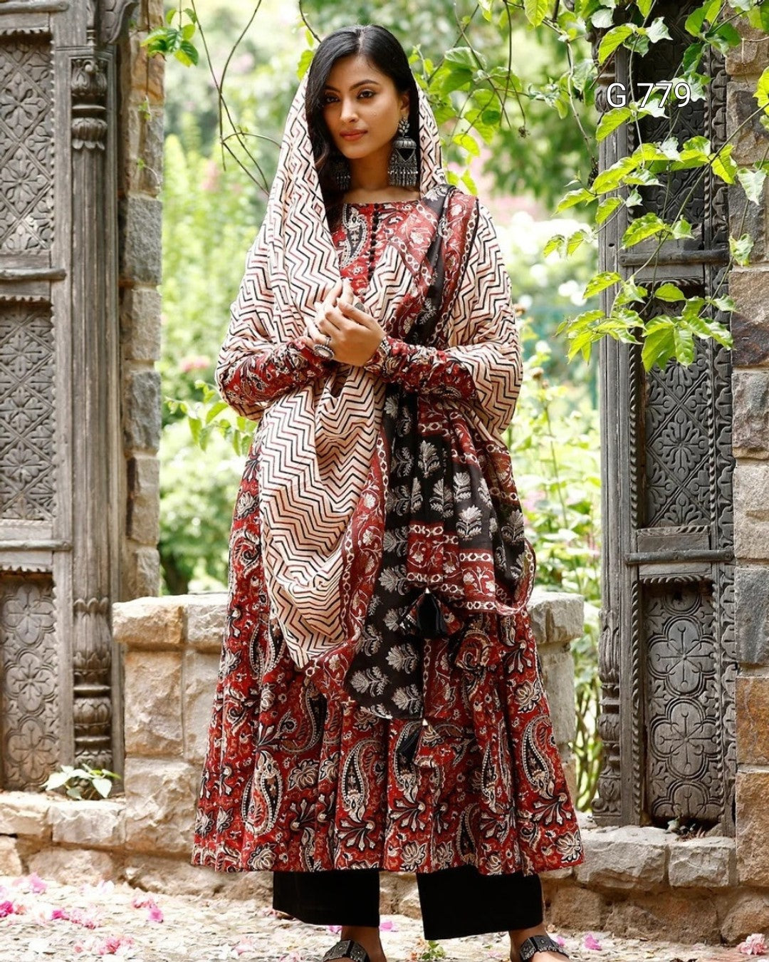 Woman wearing a patterned dress and headscarf in an outdoor setting with stone architecture and greenery.