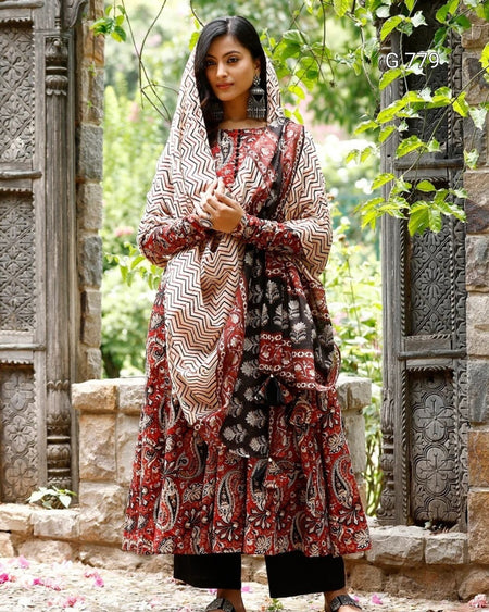 Woman wearing a patterned dress and headscarf in an outdoor setting with stone architecture and greenery.
