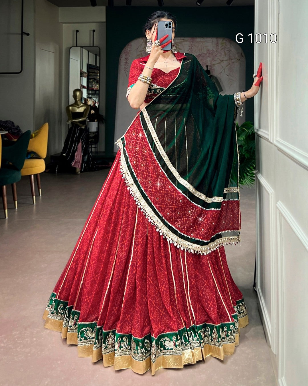 A woman takes a mirror selfie wearing a traditional Indian lehenga choli set. The outfit features a deep red flared skirt (lehenga) with green and gold borders, a matching red sleeveless blouse, and a dark green dupatta with red and gold embroidered details.