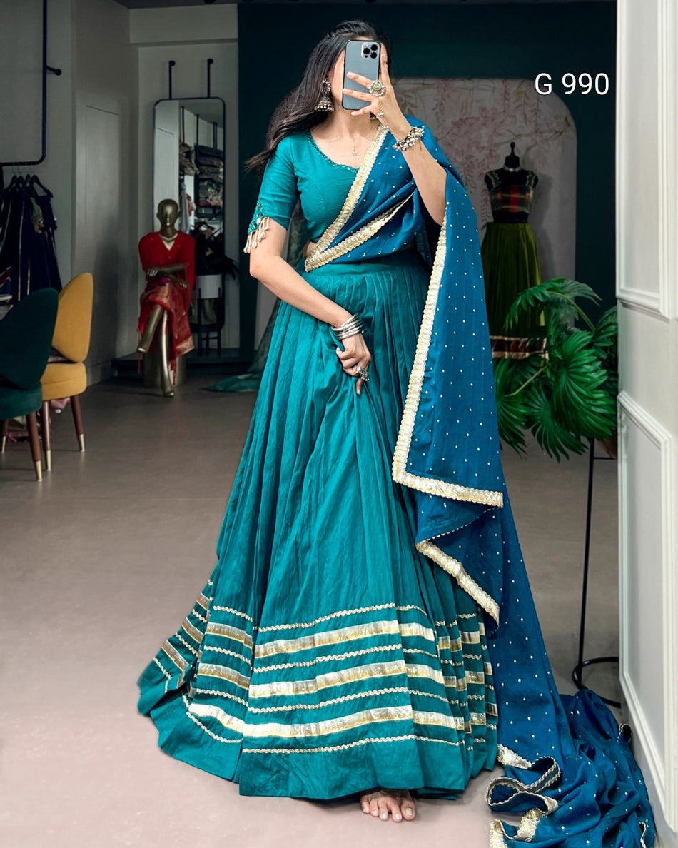 A woman takes a mirror selfie in a boutique wearing a teal blue traditional Indian dress with a matching dupatta. The dress features gold zari stripes along the hem and sleeves, and the dupatta has gold polka dots and border detailing.