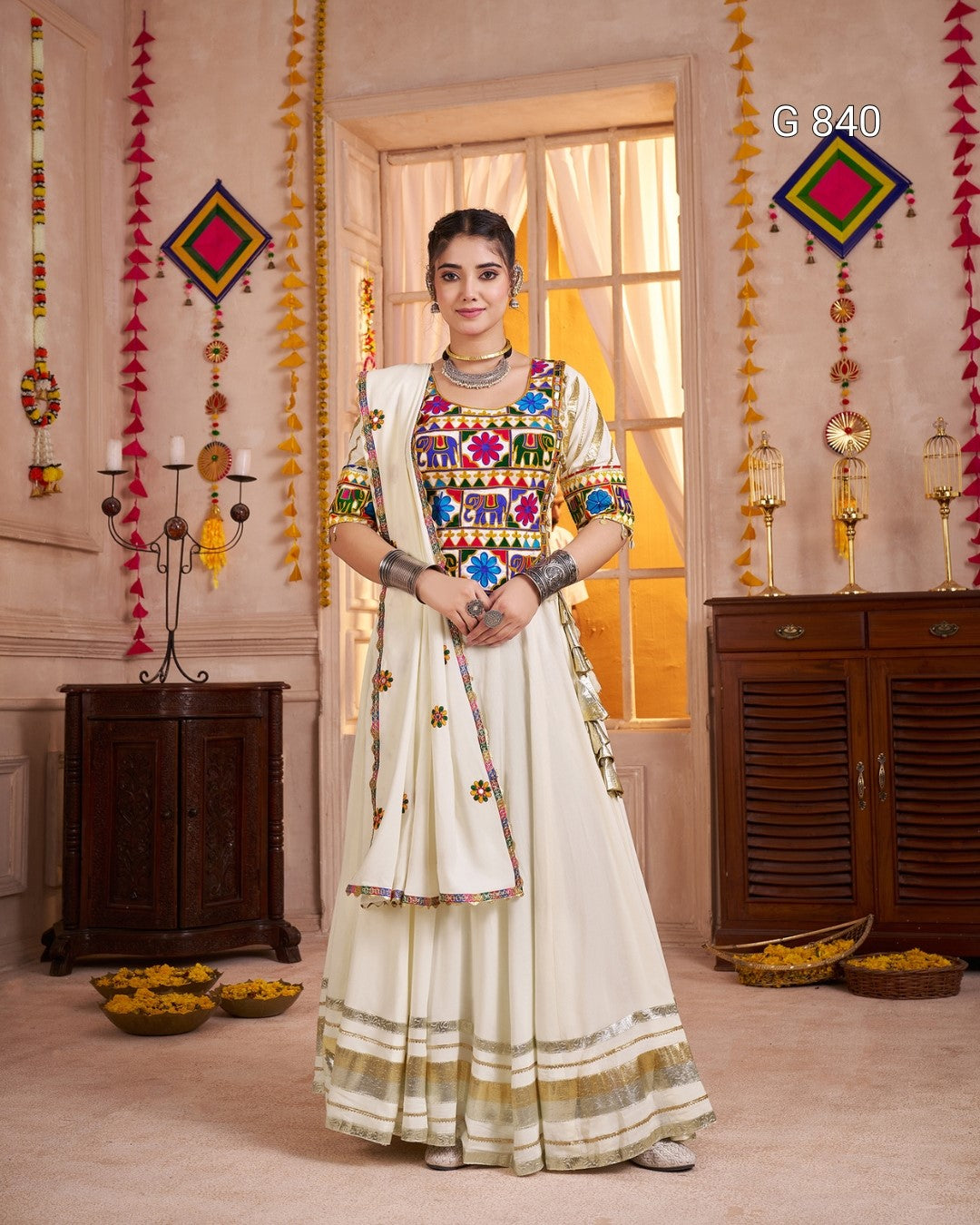 Woman in a colorful traditional outfit standing in a decorated room with hanging decorations and a window.