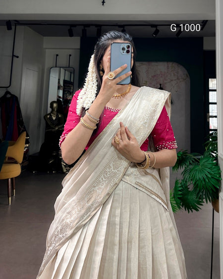 A woman takes a mirror selfie wearing a cream-colored saree with a pink embroidered blouse, gold jewelry, and a white floral hair ornament. The background shows a boutique interior with clothing racks and a plant.