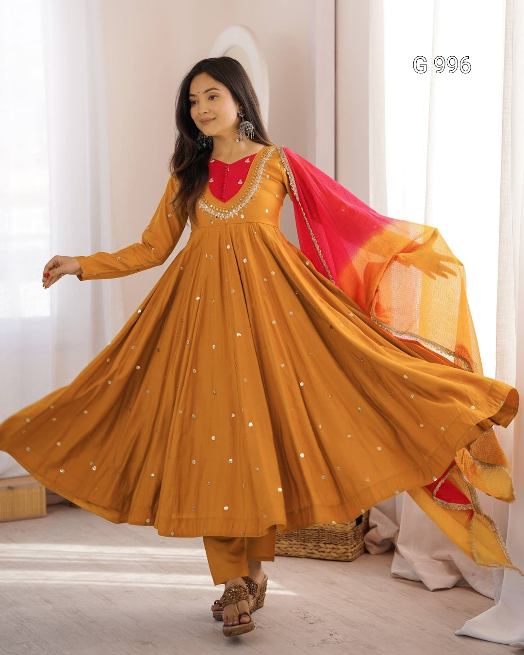 A woman modeling a mustard yellow Anarkali gown with a red embroidered neckline, gold embellishments, matching trousers, and a red and orange dupatta, captured in a bright indoor setting.