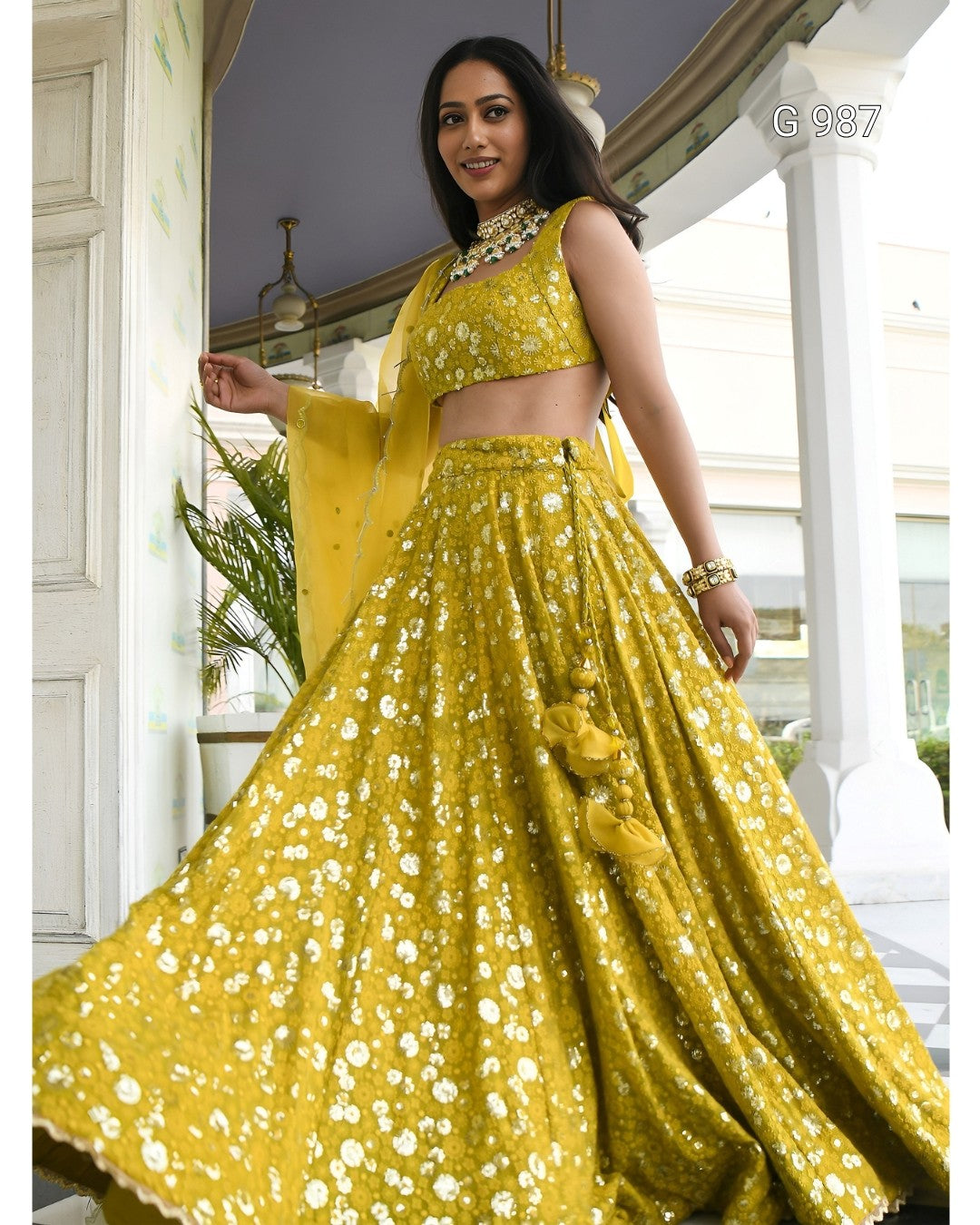 A woman models a bright yellow embellished lehenga choli set with a matching dupatta in an outdoor architectural setting. The outfit features intricate white floral embroidery and mirror work, with a matching yellow clutch.