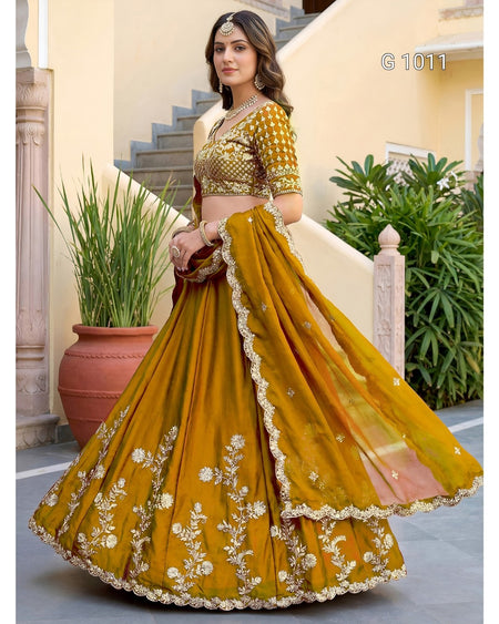 A woman models a traditional Indian lehenga choli suit in mustard yellow with gold and white floral embroidery. The set includes a heavily embroidered choli, a voluminous flared skirt (lehenga), and a matching dupatta, shown in an outdoor courtyard setting.