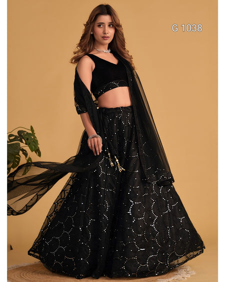 A woman models a black traditional outfit consisting of a sleeveless crop top, a flared skirt with silver embellishments, and a sheer black dupatta, set against a solid tan background.