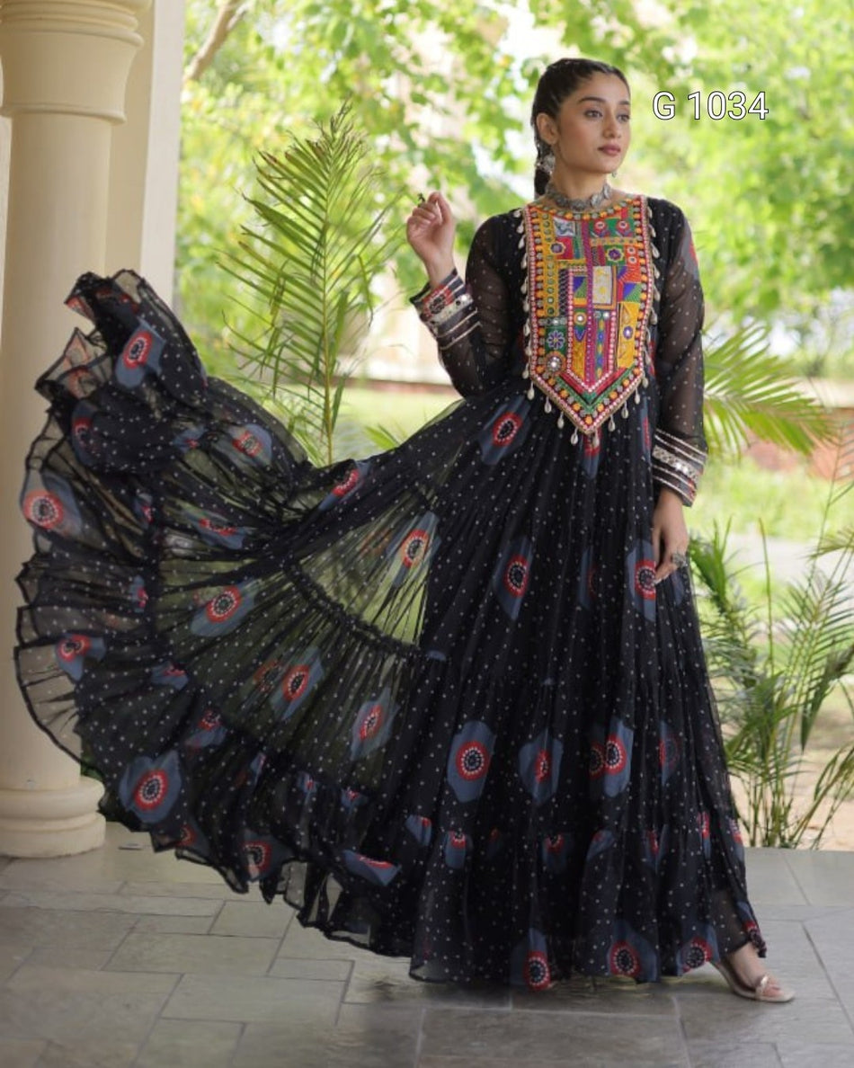 A woman modeling a black long-sleeve Anarkali gown with a heavily embroidered multicolor neckline and a flowing sheer black skirt decorated with small motifs and circular patterns, set against a garden background with a pillar.
