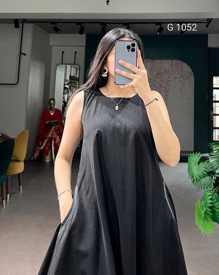 A woman takes a mirror selfie wearing a sleeveless black A-line dress with thin green piping details along the side seams. The background shows a boutique interior with a mannequin in a red dress, a yellow chair, and a green plant.
