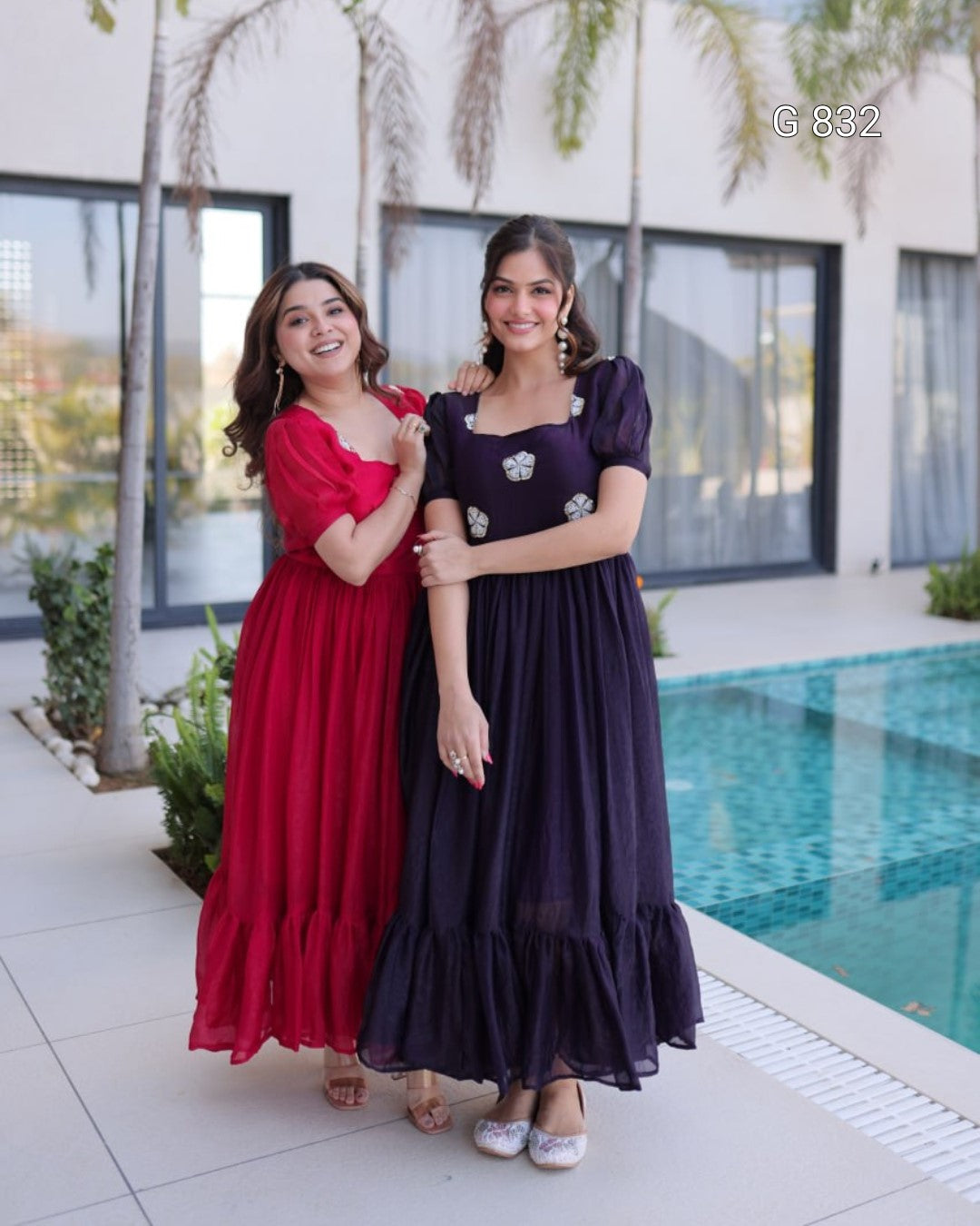 Two women in long dresses standing by a pool with a building in the background.