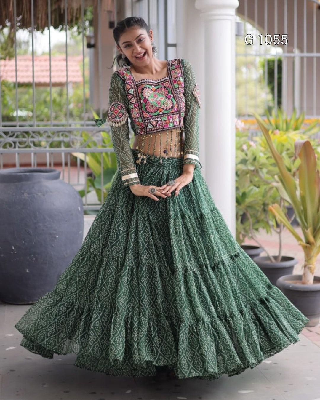 A woman modeling a traditional Indian ethnic outfit featuring a green patterned skirt (lehenga) and a matching embroidered blouse with a beige waist belt, set against a porch background with potted plants.